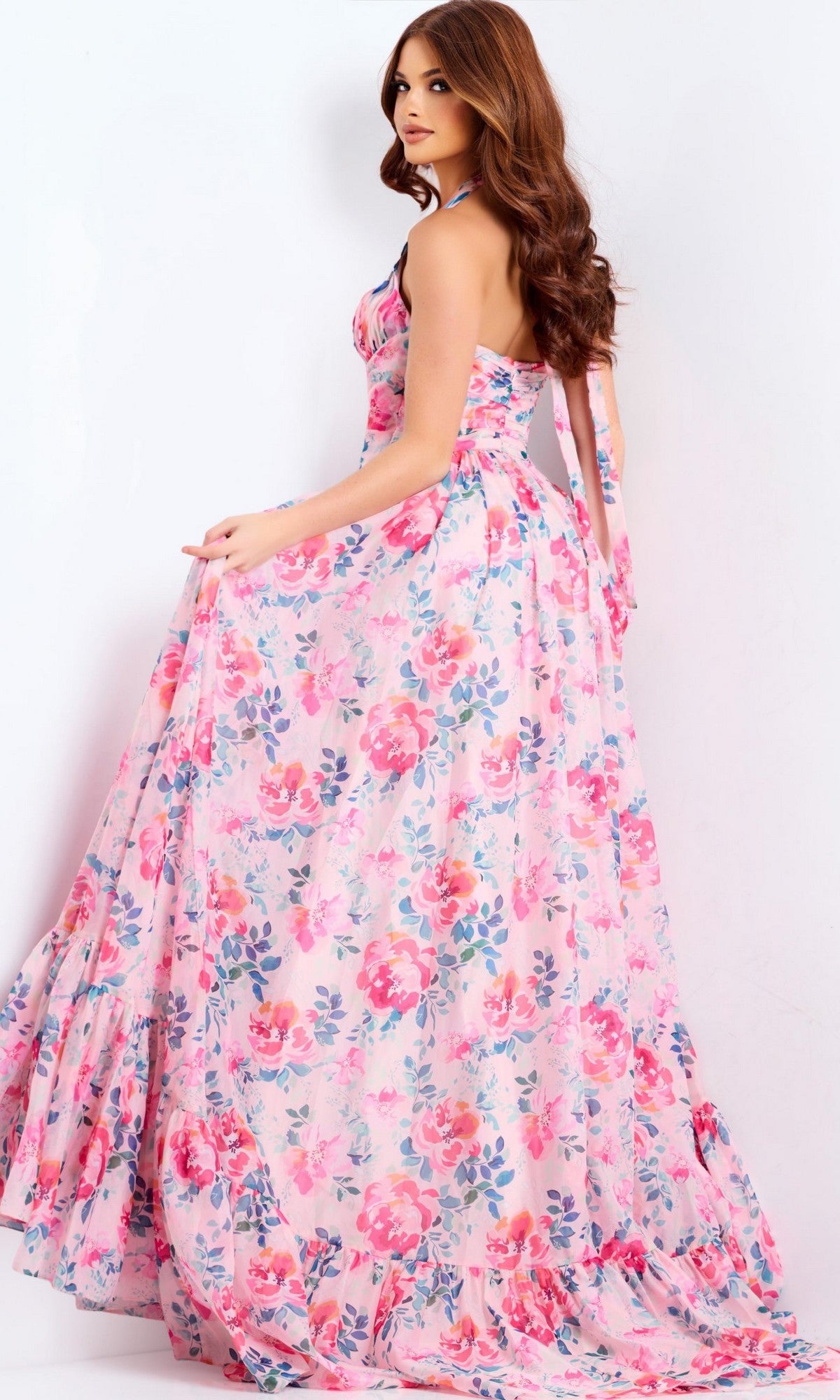 Woman in a long, sleeveless pink floral Jovani 48469 prom dress stands against a white background, looking over her shoulder and holding the skirt.